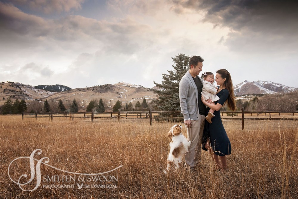 family of three and puppy king charles spaniel hugging and smiling at sunset with mountains behind them