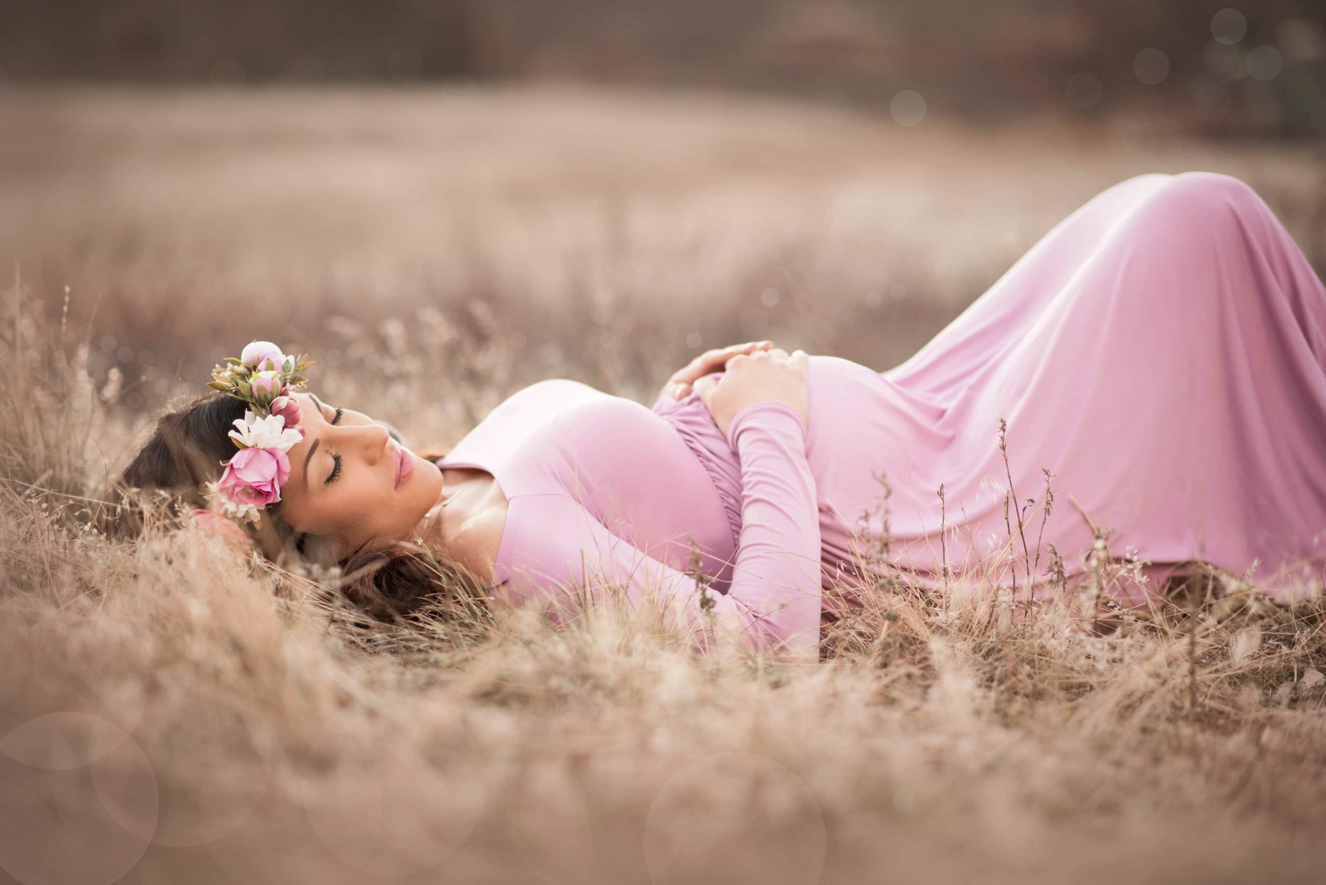 pregnant woman laying on her back in pink maternity gown with a flower crown and hands on her belly in golden field