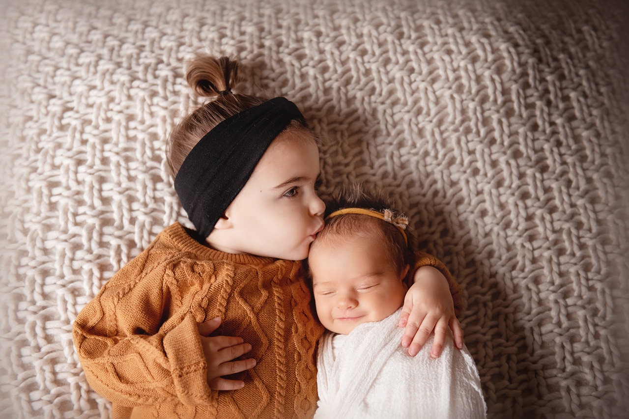 big sister kissing newborn sister smiling with mustard sweater on white blanket (boulder photographer)