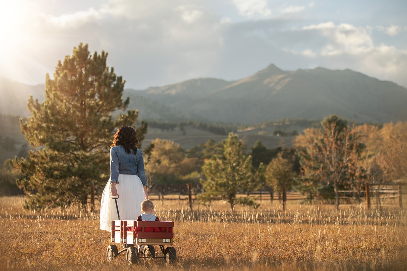 mom walking baby boy in golden field with mountain view in red wagon - boulder newborn photographer