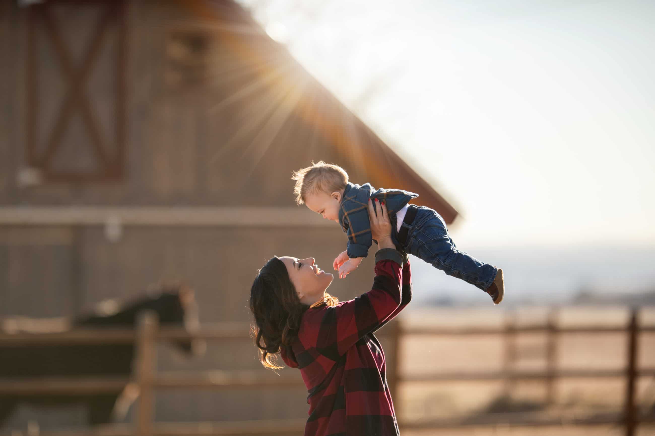 mom holding baby boy up into the air with sun flare and barn and horse - boulder newborn photographer