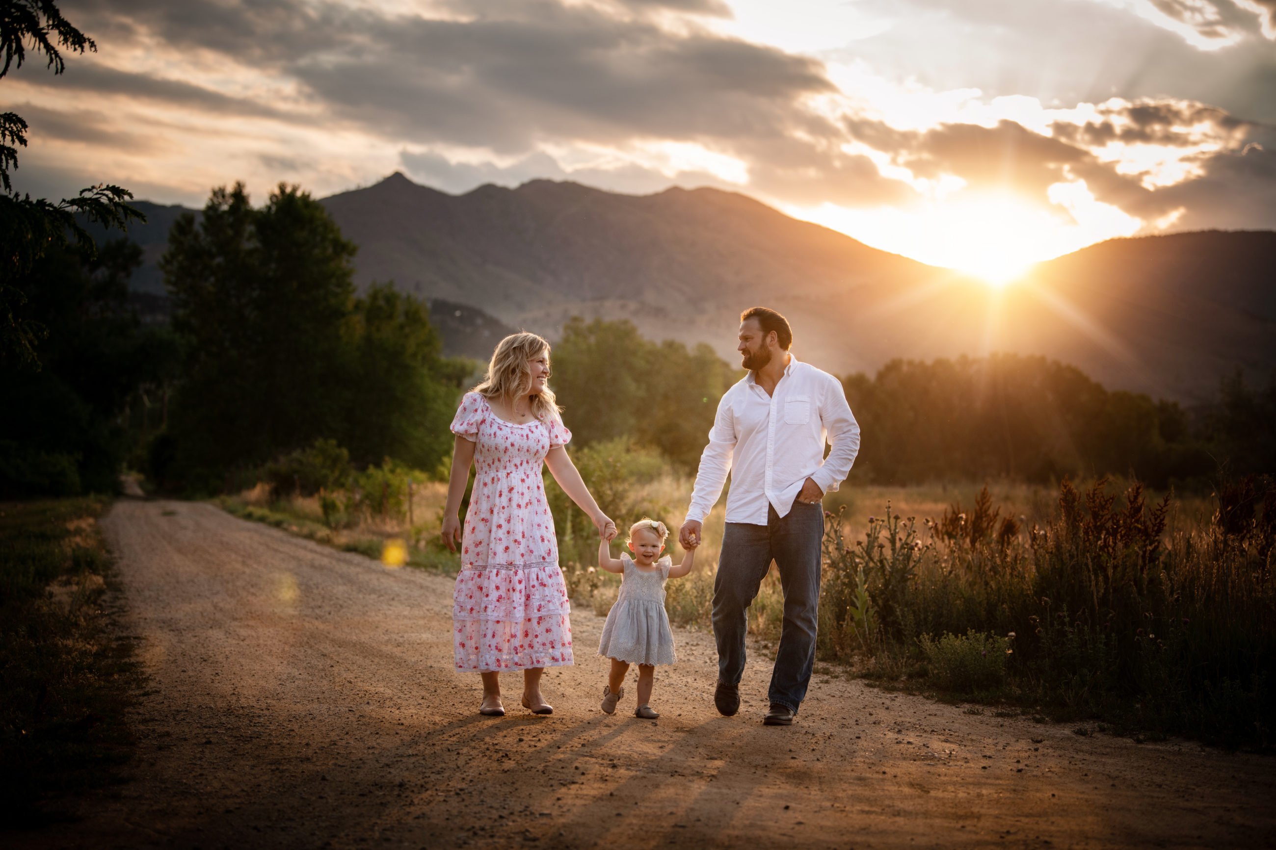 mom dad and baby girl walking down dirt road with mountains in background sun flare and pink dress